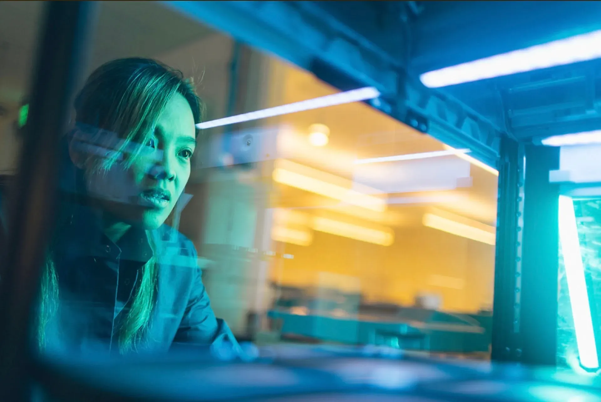 A woman working closely with sscientific equipment in a high-tech facility illuminated by cool blue and warm ambient lighting.